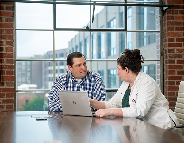 A man and woman sit at a conference table in front of a large window and talk as the woman points to a laptop screen