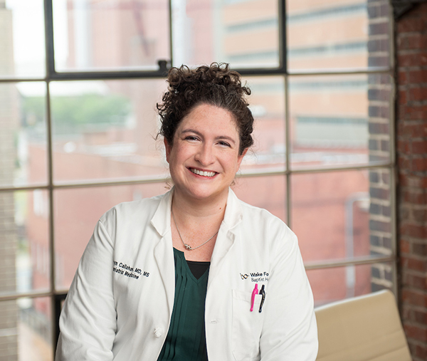 Woman with curly hair in a bun sits in front of a large window and smiles at camera