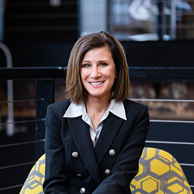 A woman in a dark blue double-breasted jacket and white shirt with dark shoulder-length hair smiles at the camera