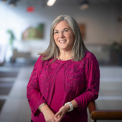 A woman in fuschia with gray hair that falls below her shoulders smiles at the camera