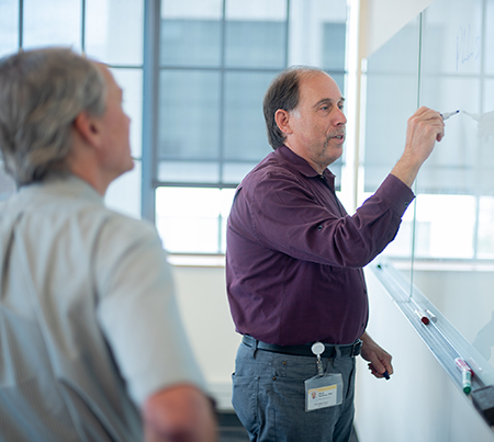 Mark Wolfson, PhD, professor of social sciences and health policy, stands at a dry-erase board and makes notes while talking with colleague