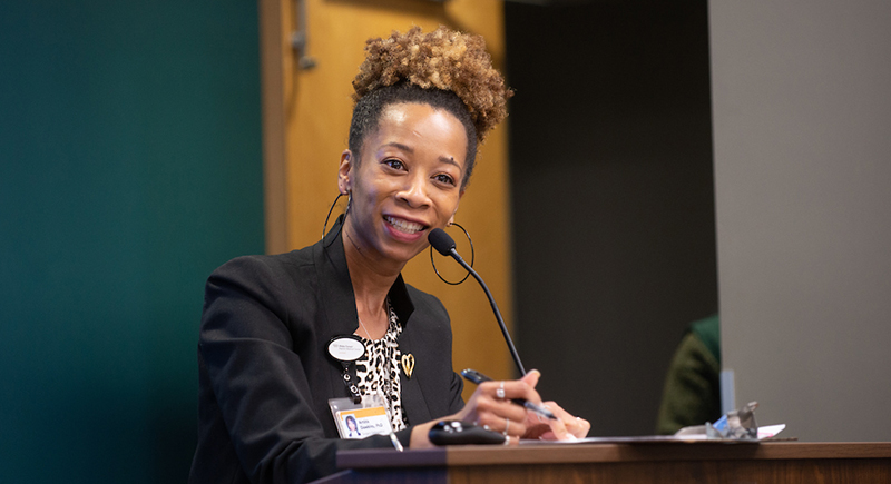 An African American woman with hair up in a high ponytail, speaks as she stands at a podium