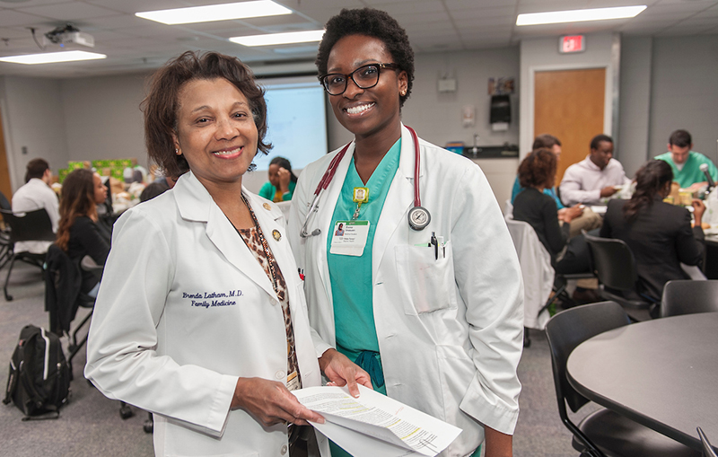 Two African American women wearing white coats stand together and smile for the camera