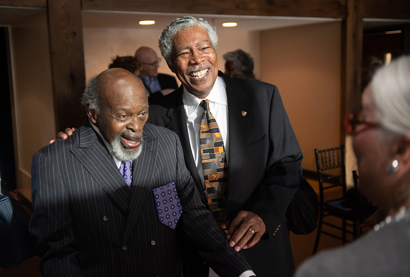 Two older African American men, wearing suits, stand next to each other and smile for the camera