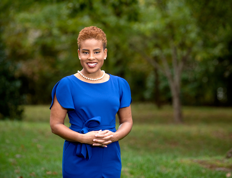 An African American woman in a blue dress with short, curly, reddish hair stands outside with blurred green vegetation in the background