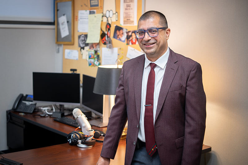 A man in a suit standing next to a desk with two computer monitors on it