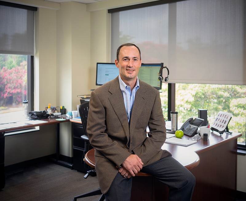 A man in a brown jacket and blue button-down shirt sits on the edge of a desk in an office and smiles at the camera