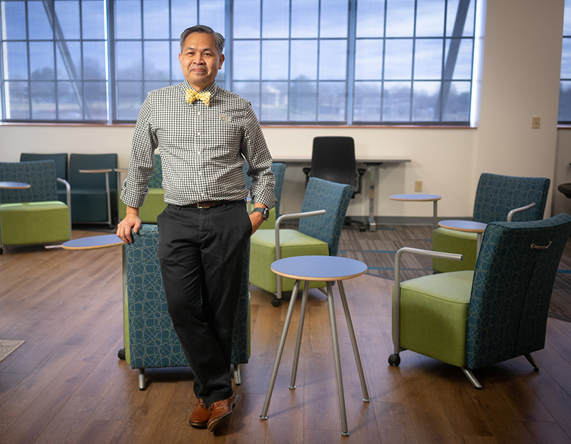 An Asian man in checked button-down shirt and bow tie stands in a lounge/study area with blue and green chairs