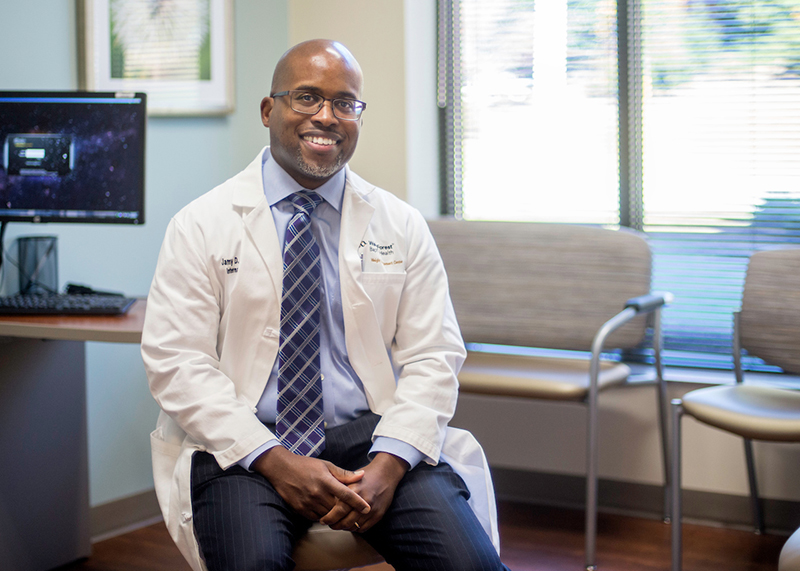 An African American man wearing glasses, shirt and tie, and a white coat smiles as he sits in an exam room