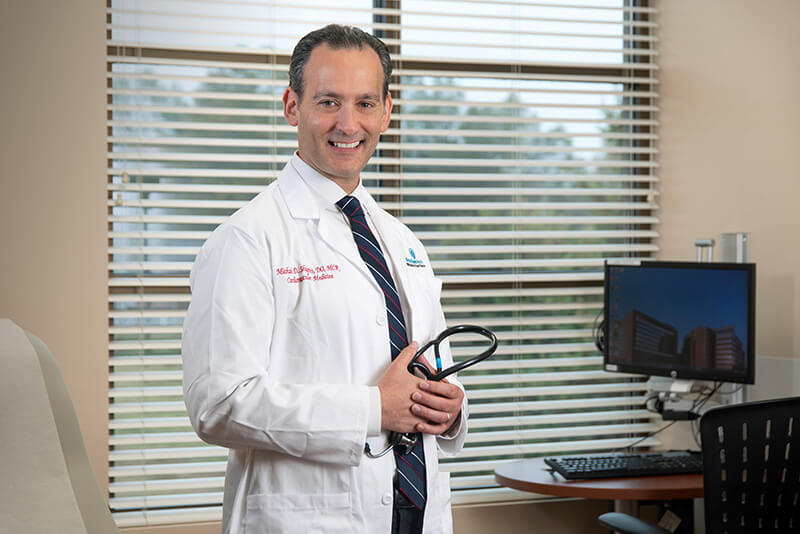 An older man wearing a lab coat and holding a stethoscope while smiling at the camera.