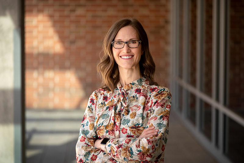 A white woman with shoulder-length brown hair, wearing glasses and a patterned blouse, stands in a sunlit corridor with arms crossed and smiles