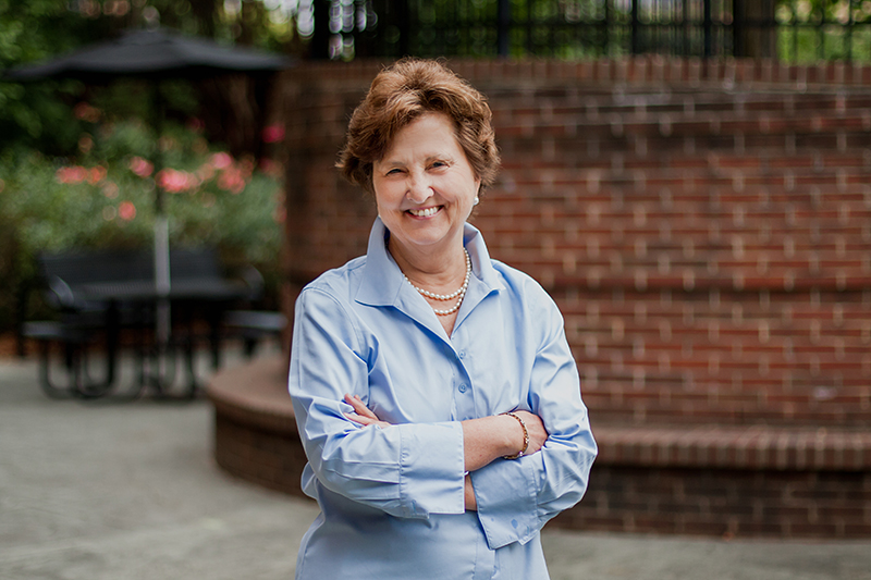 An older white woman in a blue button-down shirt smiles and stand with her arms folded