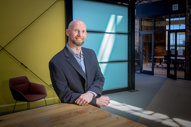 A bearded man in a blue shirt and navy jacket leans against a conference room table and smiles