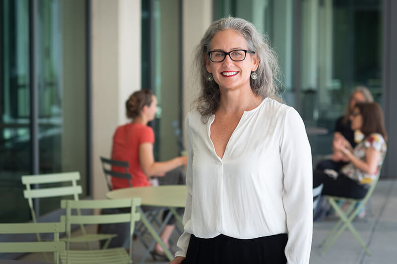 A woman with long gray hair and glasses, wearing a white shirt and smiling at the camera.