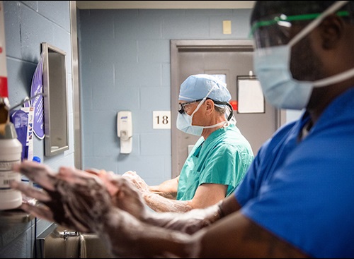 An African American man in blue scrubs and a white man in green scrubs lather up their hands before going into surgery. Both men have head coverings and surgical masks on