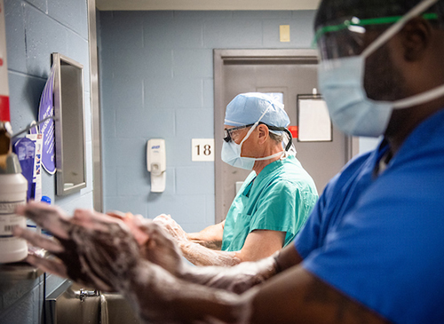 An African American man in blue scrubs and a white man in green scrubs lather up their hands before going into surgery. Both men have head coverings and surgical masks on