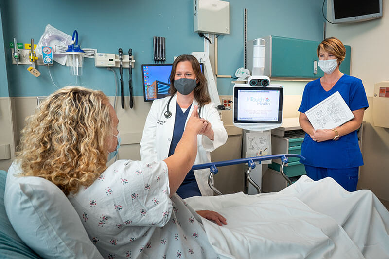 Two doctors, one in a lab coat and one in blue scrubs talk to a patient in a hospital bed.