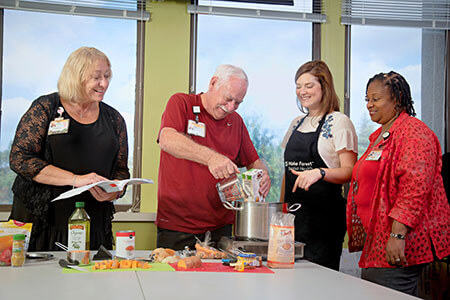 Two older people, one man and one woman, taking part in a cooking class as two women staff members observe,