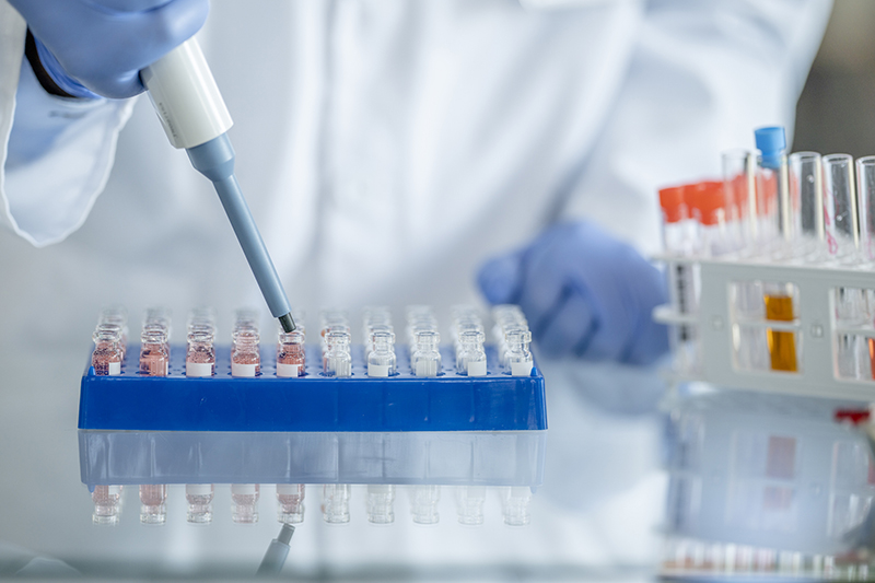 A person wearing white PPE and blue gloves uses a pipette to put liquid into test tubes, which are lined up in a blue tray and sitting on a reflective counter