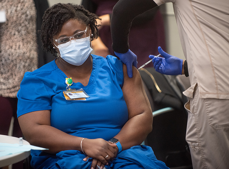 An African American woman in blue scrubs and wearing glasses and a facial mask receives an injection in her left arm from a person in tan scrubs