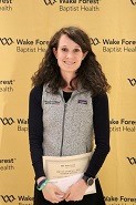 A young brunette woman stands in front of a Wake Forest Baptist Health gold background and smiles