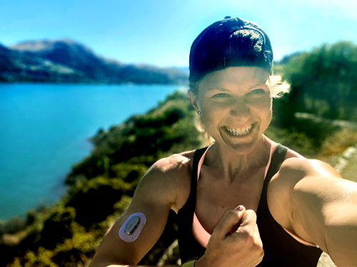 Blonde, fit woman wearing a hat and continuous glucose monitor on her right arm, poses for a selfie in front of a blue body of water