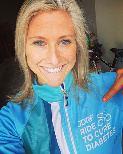 Blonde young woman with blue eyes and tan smiles and points to the JDRF Ride to Cure Diabetes logo on her light blue jacket