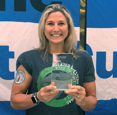 Young, blonde woman wearing a blue and green cycling shirt smiles as she holds up a trophy