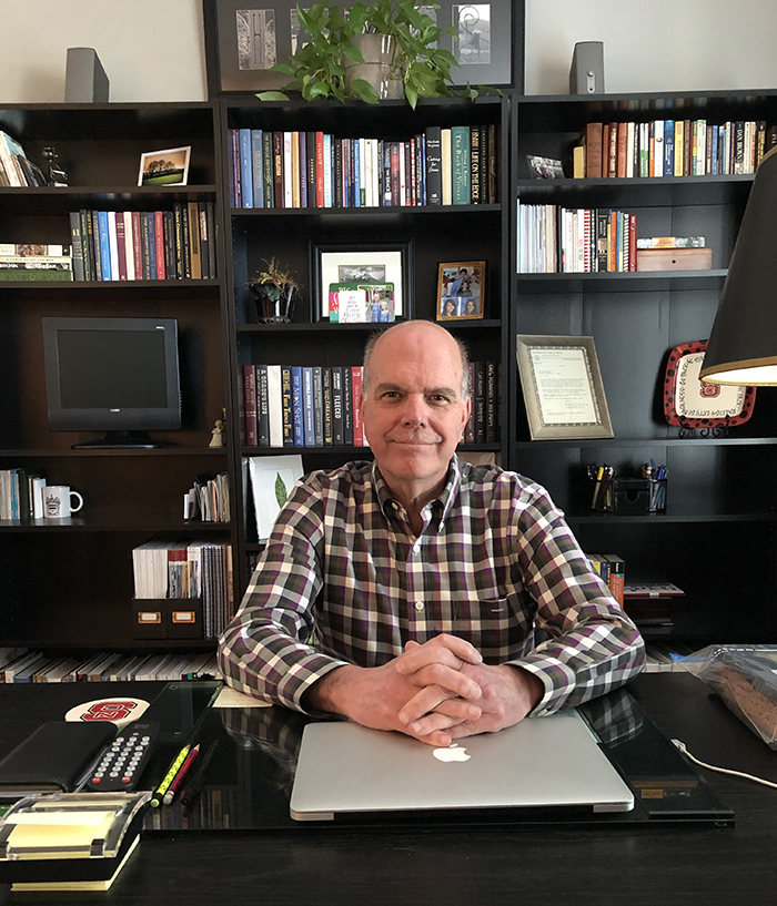 An older, balding man sits at a dark wood desk with bookshelves full of books behind him