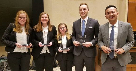 Three young women and two young men, all in business attire, stand in a row and smile for the camera