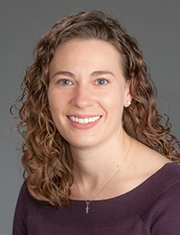 Head shot of young woman with curly brown hair, smiling in front of a grey background