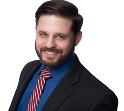A man with brown hair, a beard and mustache wearing a suit and tie smiles