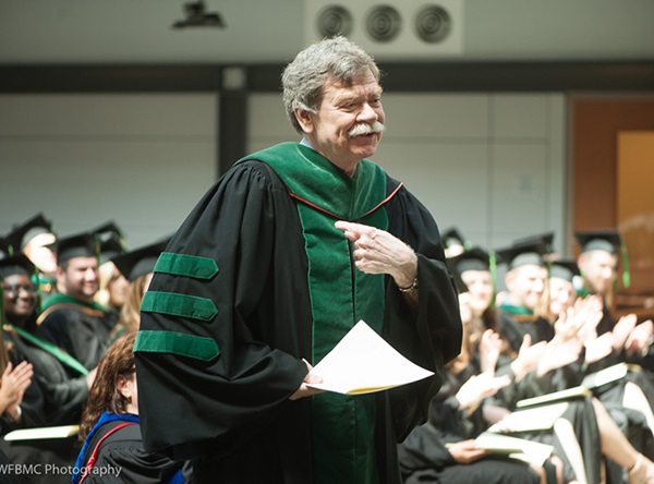 A man with grey hair and a grey mustache, wearing a black commencement gown with green stole, holds papers and smiles as he walks past graduating students