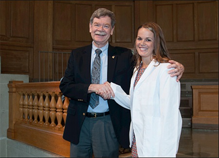 A grey-haired man in a jacket and tie smiles at the camera as he shakes the hand of a young lady wearing a white coat