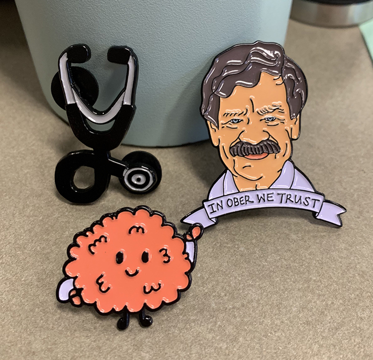 A trio of pins - a stethoscope, an orange fluffy ball with eyes, and Dr. Pat Ober's face with 'In Ober We Trust' beneath it - sits on a desk