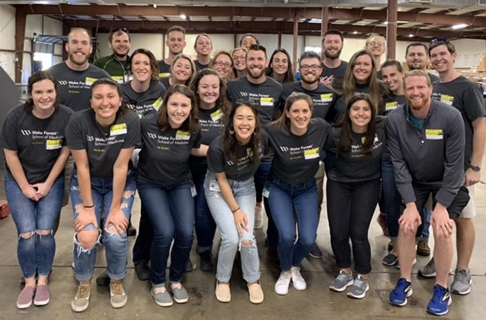 A large group of young people wearing black Wake Forest School of Medicine PA program shirts stand and pose in a food warehouse