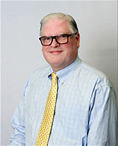 A blond man wearing dark-rimmed glasses, a light blue button-down shirt and yellow tie smiles at the camera