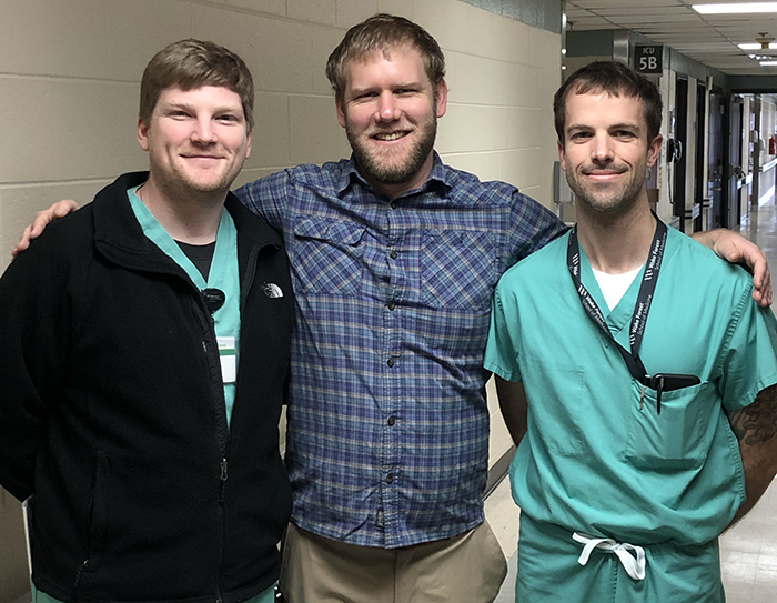 Three young men, one in scrubs and two in regular clothes, stand in a hallway and smile for the camera