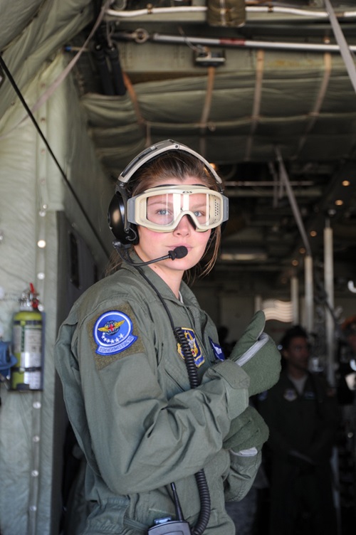 A young woman in military flight uniform, helmet and goggles stands in the hold of an airplane