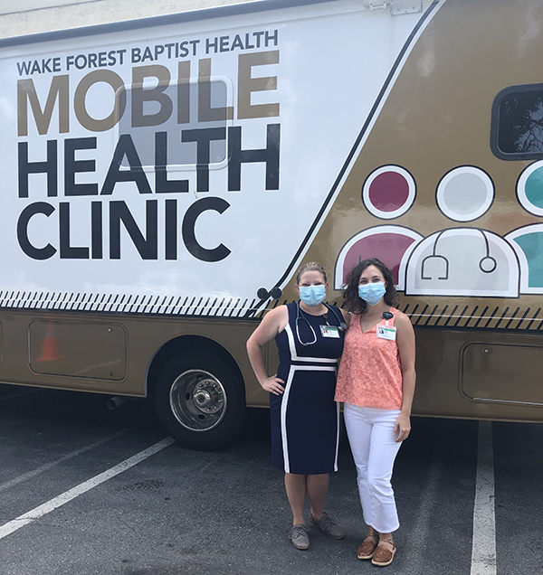 Two women wearing blue face masks stand outside the Wake Forest Baptist Health Mobile Health Clinic