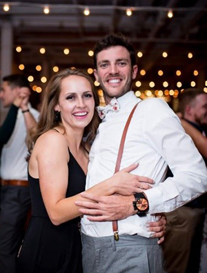 A man and woman with arms around each other smile at the camera with lights and people dancing in the background