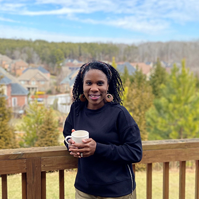 A Black woman stands on a balcony with a cup of coffee, houses and a blue sky behind her