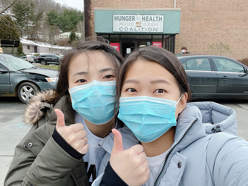 Two young women wearing blue face masks and jackets give a thumbs-up gesture in front of a brick building
