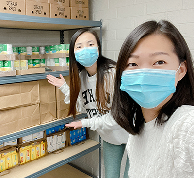 Two young women wearing blue face masks gesture toward neatly arranged pantry shelves