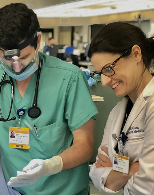 Preceptor Jennifer Nelson, MD, wearing her white coat laughs as she watches a student wearing green scrubs and a face mask perform a procedure