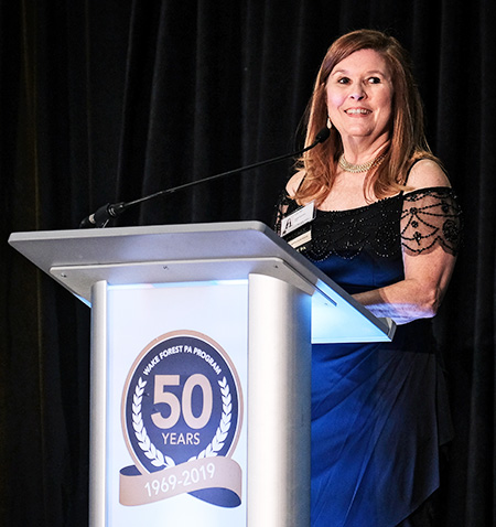 Gail Curtis, associate professor and chair of the Department of PA Studies, wears a black dress with lace sleeves as she speaks at the podium for Wake PA's 50th Anniversary