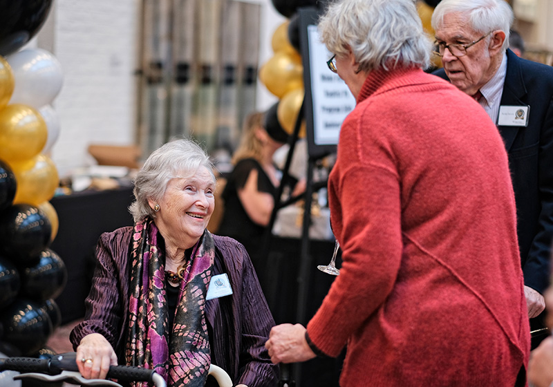 An older woman sits and smiles at another older woman wearing a red jacket; an older man stands in the background