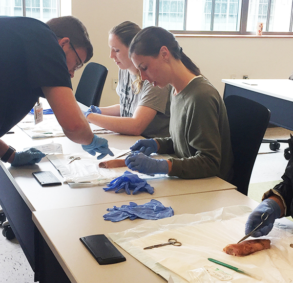 Female student seated at table in room with lots of natural light looks at object she is cutting as instructor points out something