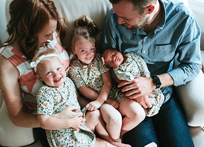 Looking down from above, a white family (man, woman, two girls and newborn) sit on a couch and smile at each other