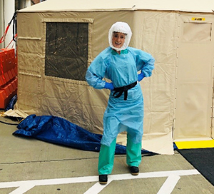 A person in green scrub pants, blue gown and hazmat hood stands with hands on hips outside a beige tent